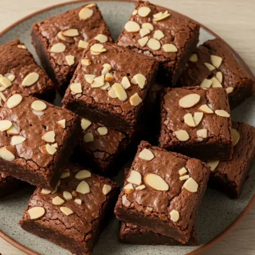 almond butter brownies on a rustic plate with almonds and chocolate chunks on a light wooden table.