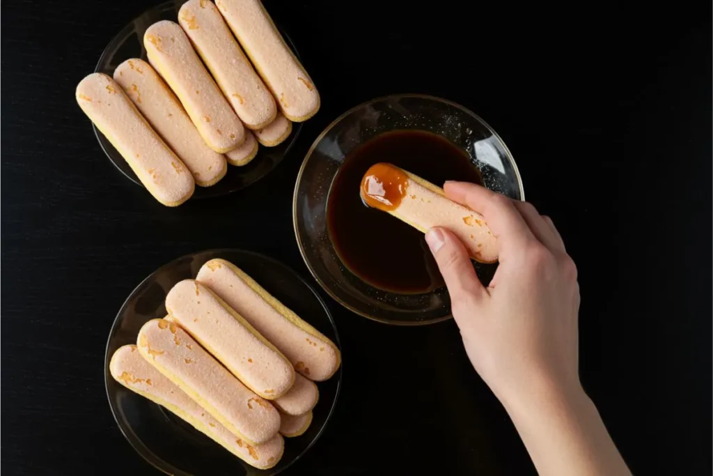 Top-down view of ladyfingers being dipped into coffee on a black table during tiramisu preparation.