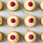 Mini Egg Cheesecakes arranged on white marble surface with whipped cream and raspberry toppings, showing golden graham cracker crusts and creamy texture