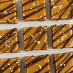 Golden brown salted caramel protein bars with dark chocolate drizzle and sea salt crystals, arranged on a white plate