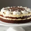 A homemade Brownie Ice Cream Cake on a white cake stand, featuring layers of fudgy brownie, creamy vanilla ice cream, and rich chocolate ganache, topped with fluffy whipped cream and delicate chocolate shavings.