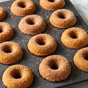 Homemade mini apple cider donuts in donut pan with golden-brown baked rings coated in sparkling cinnamon-sugar on white marble surface