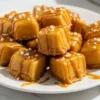 Homemade mini caramel apple bites on wooden board with glossy golden caramel coating and flaky sea salt sprinkles on white marble surface