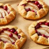 Homemade mini pear and cranberry galettes on parchment-lined baking sheet with rustic golden pastry and ruby-red cranberries on marble surface