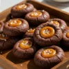 Mini chocolate-salted caramel cookies on rustic wooden tray with fudgy chocolate exterior, glossy caramel centers, and flaky sea salt crystals