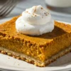 Single pumpkin pie bar slice on white plate showing golden crust layer beneath creamy spiced pumpkin custard filling with whipped cream topping