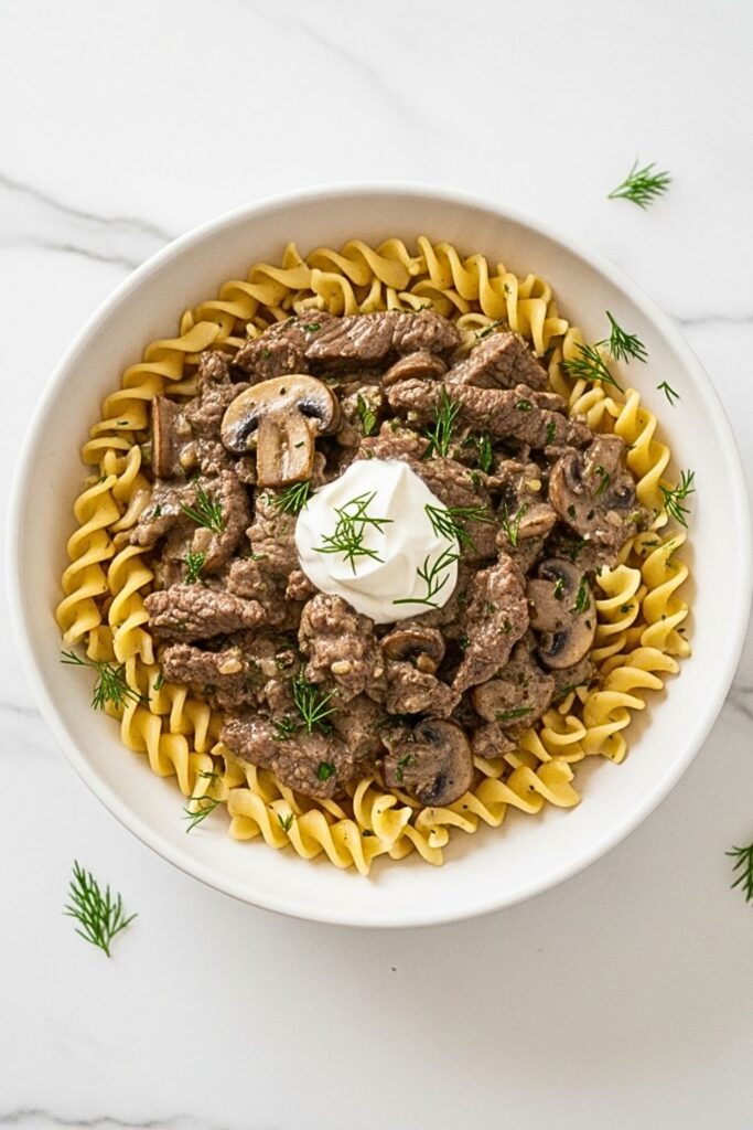 beef stroganoff over noodles in a white bowl on a clean white marbled table.