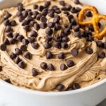 A close-up of a white bowl filled with creamy chocolate chip cookie dough dip, topped with mini chocolate chips and a salty pretzel.