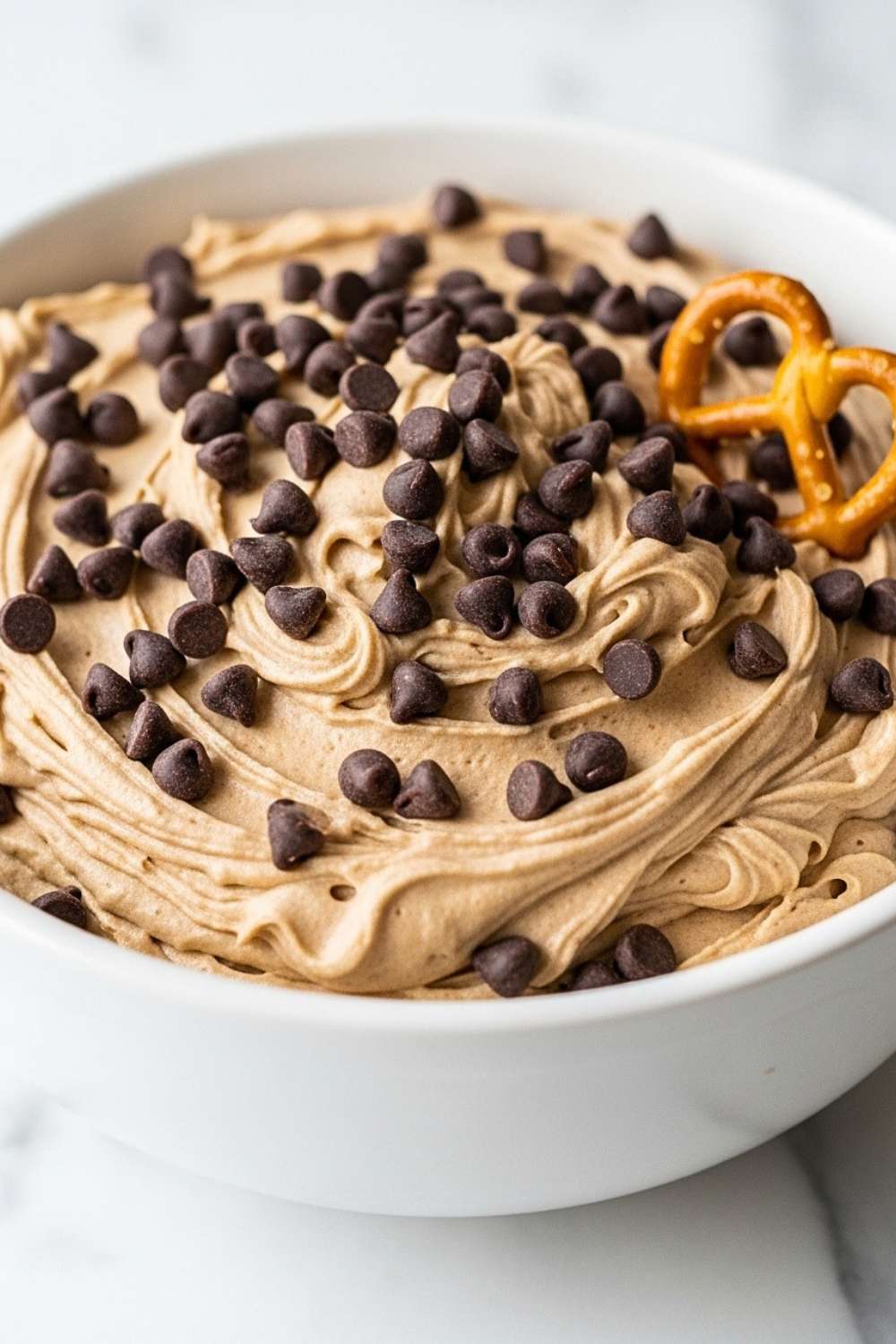 A close-up of a white bowl filled with creamy chocolate chip cookie dough dip, topped with mini chocolate chips and a salty pretzel.